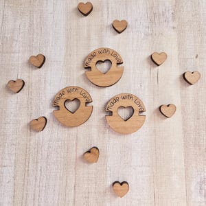 May include: Wooden heart-shaped embellishments and three circular wooden pieces with a heart cutout and the words "made with Love" engraved. The hearts and circles are arranged on a light-colored, wood-grain surface.