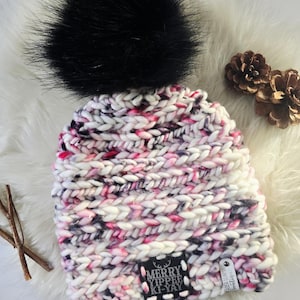 May include: A chunky knit beanie featuring a variegated yarn pattern in white, pink, and black, topped with a large black faux fur pom-pom. A black square patch on the front displays the white text MERRY YIPPEE KI-YAY with a small deer icon.