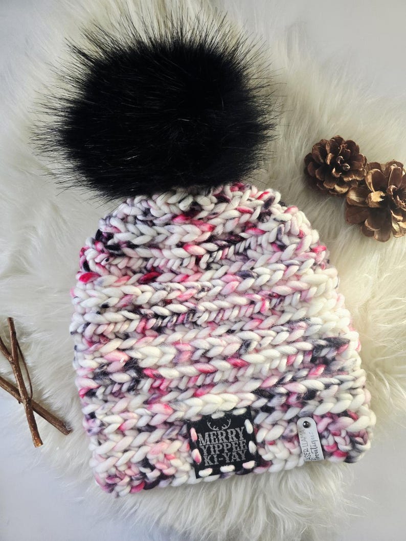 May include: A chunky knit beanie featuring a variegated yarn pattern in white, pink, and black, topped with a large black faux fur pom-pom. A black square patch on the front displays the white text MERRY YIPPEE KI-YAY with a small deer icon.