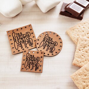May include: Three wooden plaques with the text "I'm a happy camper" in a handwritten font. The plaques are in different shapes and sizes, with a square, a circle, and a smaller square. The plaques are on a white wooden surface.