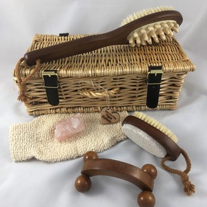 Puede incluir: Una cesta de mimbre llena de productos de baño y cuerpo, que incluyen un cepillo de espalda de madera, un cepillo de cuerpo de madera, un masajeador de espalda de madera, una esponja vegetal y un cristal rosa. La cesta tiene una etiqueta que dice "With Love from Vegan Embrace".