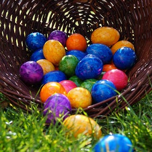 Può includere: Un cesto di vimini traboccante di uova di Pasqua colorate. Le uova sono dipinte in tonalità di blu, verde, giallo, viola e rosso. Alcune delle uova hanno un effetto marmorizzato.