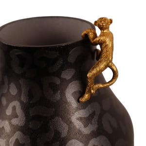 Op de afbeelding: Een zwarte vaas met een luipaardprint en een goudkleurige aap die over de rand klimt. De vaas heeft een brede basis en een smalle hals, en de aap is gedetailleerd met vacht en gezichtskenmerken.