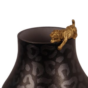 Può includere: Un vaso nero con una figura di leopardo dorata sul bordo. Il vaso ha una superficie scura e strutturata con un motivo maculato. La figura è dettagliata e aggiunge un tocco decorativo.