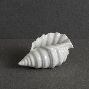 Może przedstawiać: Biała ceramiczna rzeźba w kształcie muszli z błyszczącym wykończeniem. Obiekt posiada zwężający się, spiralny czubek na jednym końcu oraz szeroki, karbowany otwór na drugim, naśladujący naturalne grzbiety i organiczne krzywizny muszli.