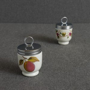 May include: Two white porcelain egg coddlers with silver-toned metal lids featuring circular ring handles. The front coddler displays a single plum with green leaves, while the coddler in the background features a vine pattern with grapes.