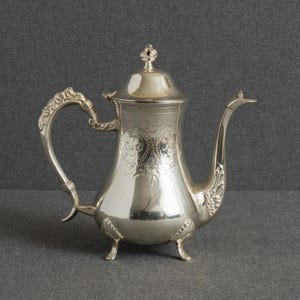 May include: A polished silver teapot featuring an ornate handle with scrollwork, a curved spout, and a lid topped with a small finial. The body is decorated with engraved floral patterns and rests on three decorative feet with shell-like details.