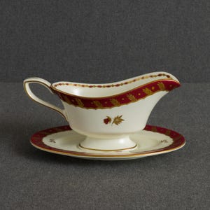 Op de afbeelding: Een crèmekleurige porseleinen sauskom met bijpassende schotel, voorzien van een donkerrode rand langs de bovenkant, geaccentueerd met gouden patronen van korenaren en kleine bloemmotieven. De sauskom heeft een gebogen handvat en een brede tuit.