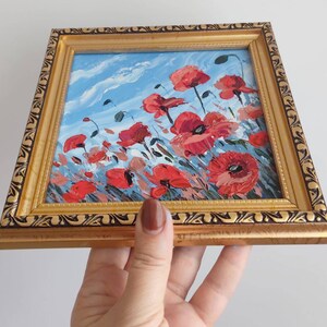 May include: A framed painting depicting a field of red poppies against a blue sky. The painting is in a gold frame with an ornate border.