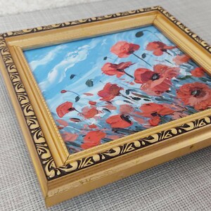 May include: A framed painting depicting a field of red poppies against a blue sky. The painting is in a gold frame with a black and gold design.