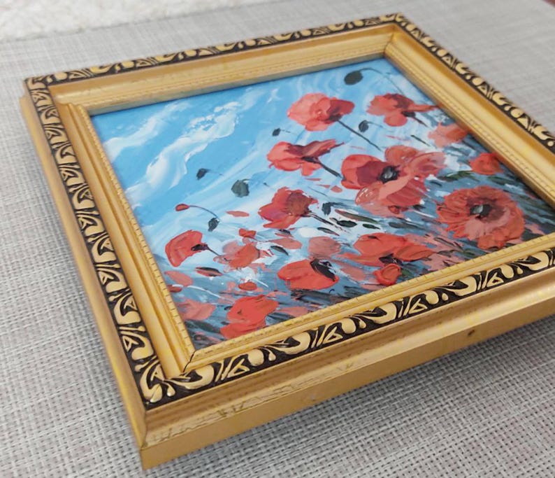 May include: A framed painting depicting a field of red poppies against a blue sky. The painting is in a gold frame with a black and gold design.