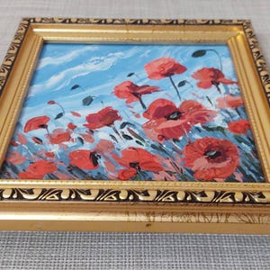 May include: A framed painting depicting a field of red poppies against a blue sky with white clouds. The painting is in a gold frame with a black and gold design.