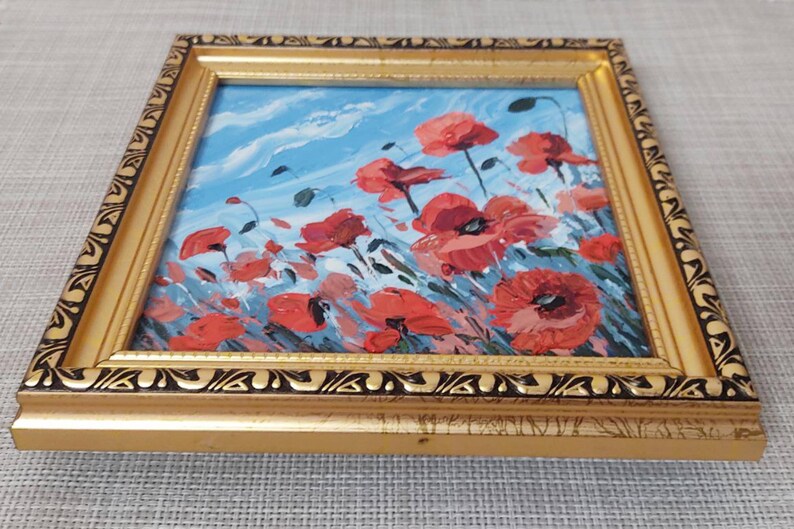 May include: A framed painting depicting a field of red poppies against a blue sky with white clouds. The painting is in a gold frame with a black and gold design.