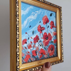 May include: A framed painting depicting a field of red poppies against a blue sky with white clouds. The frame is gold with an ornate design.