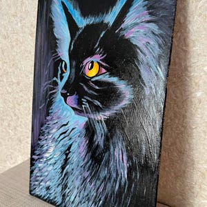 May include: A painting of a black cat with bright yellow eyes, set against a dark background. The cat's fur is painted with shades of blue and purple, creating a vibrant and abstract effect. The artist's signature, "Olya", is visible in the bottom right corner.