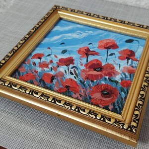 May include: A framed painting depicting a field of red poppies against a blue sky. The frame is gold with a black ornate design.