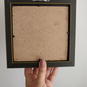May include: The back of a dark green picture frame with a brown cardboard backing. The frame has a metal hanging hook on the top.