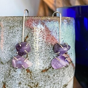 Puede incluir: Un par de pendientes de plata con tres piedras de amatista cada uno. Las piedras son de color púrpura claro y tienen una forma irregular.