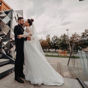 Może przedstawiać: Para młoda para pozuje do zdjęcia ślubnego przed nowoczesnym budynkiem z lustrzaną fasadą. Panna młoda ma na sobie białą suknię ślubną z długim welonem, a pan młody ma na sobie czarny garnitur. Para stoi na schodach.