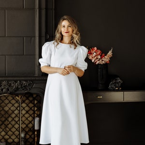 May include: A woman in a white dress with puffed sleeves stands in front of a dark fireplace. She is wearing nude heels and has her hair pulled back.