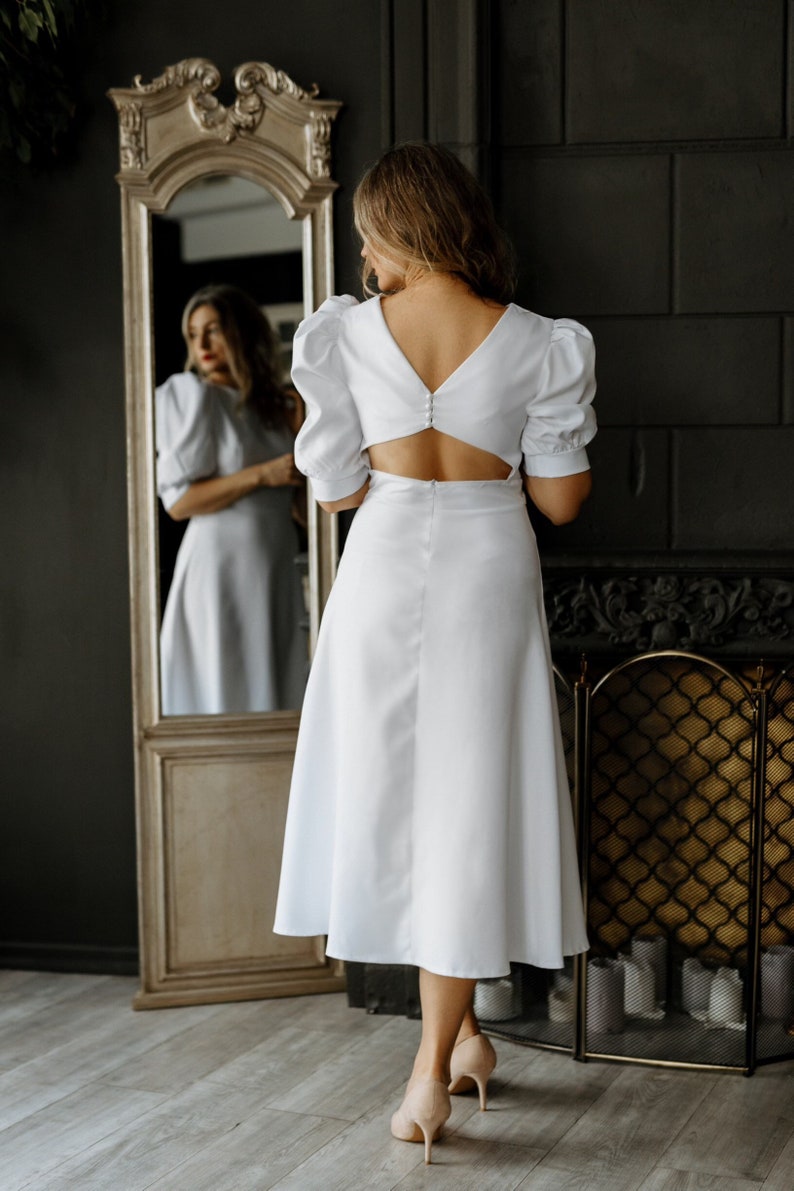 May include: A white midi dress with a low back and puff sleeves. The dress has a button closure at the back and a fitted bodice. The model is wearing the dress with nude heels.