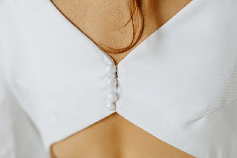 May include: White button-up shirt with a V-neckline and a row of pearl buttons down the center.