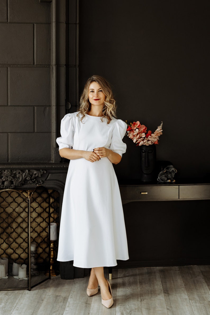 May include: A woman in a white dress with puffed sleeves stands in front of a dark fireplace. She is wearing nude heels and has her hair pulled back.