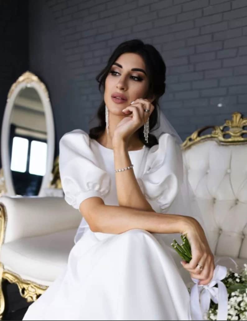 May include: A woman in a white wedding dress with a white veil and a bouquet of white flowers. She is sitting on a white chair with gold accents.