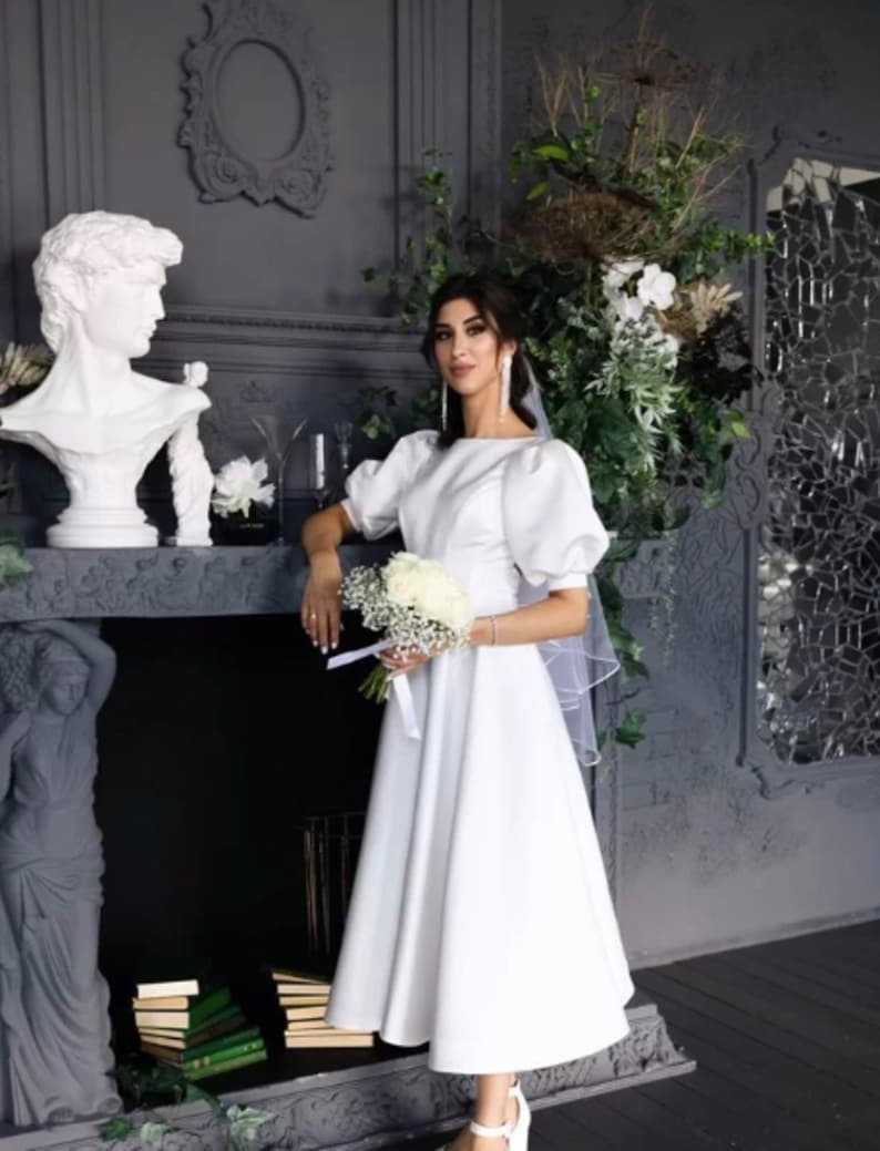 May include: A white bridal gown with a fitted bodice and a full skirt. The gown has short puff sleeves and a bateau neckline. The bride is holding a bouquet of white flowers.