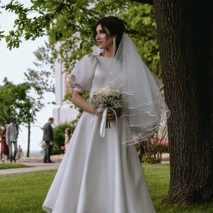May include: A bride wearing a white satin wedding dress with a full skirt and puff sleeves. She is holding a bouquet of white flowers and wearing a veil.