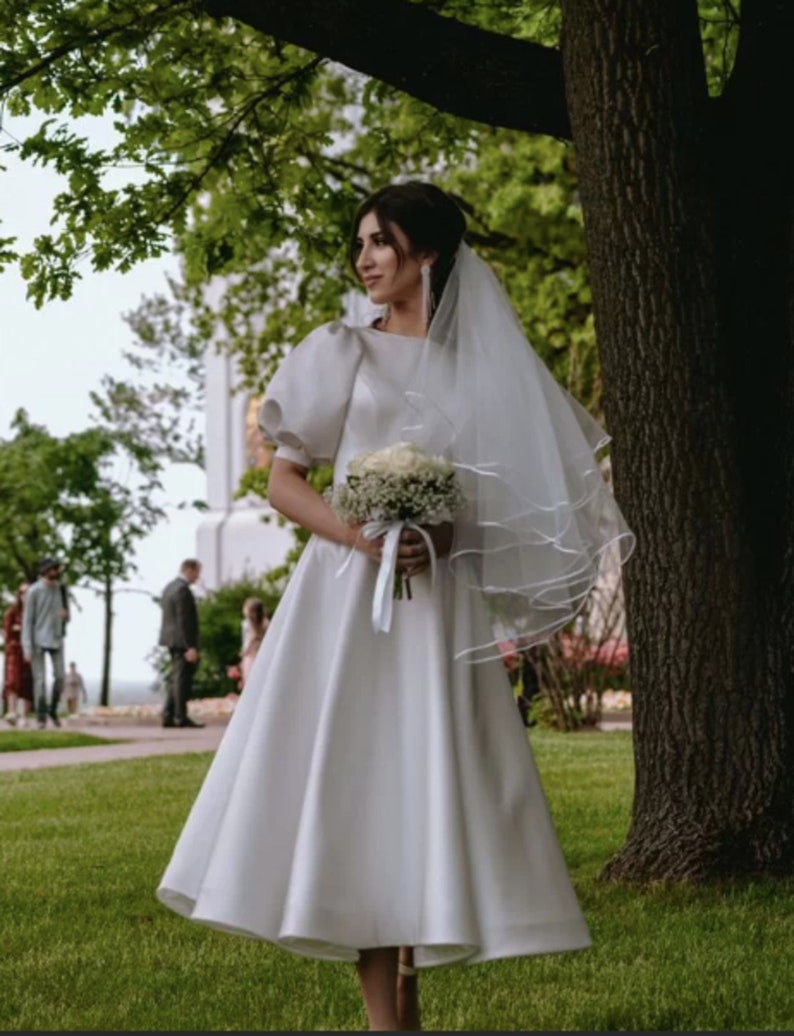 May include: A bride wearing a white satin wedding dress with a full skirt and puff sleeves. She is holding a bouquet of white flowers and wearing a veil.