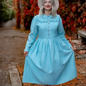 Puede incluir: Vestido azul claro con cuello redondo y mangas largas. El vestido tiene una botonadura frontal y una falda fruncida. La persona lleva un sombrero blanco y zapatillas blancas. El fondo presenta follaje rojo y un camino de ladrillos.