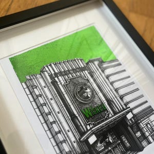 Puede incluir: Un dibujo en blanco y negro del exterior de un teatro con la palabra "Wicked" en letras verdes sobre la entrada. El dibujo est&aacute; enmarcado y tiene un fondo verde.
