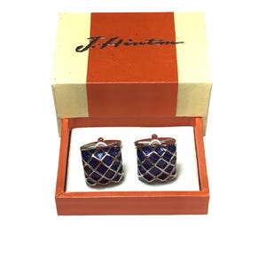 Op de afbeelding: Een paar manchetknopen in een doos. De manchetknopen zijn zilver met een blauw emaille ontwerp. De doos is oranje met de tekst "J. Hinton" op de bovenkant.