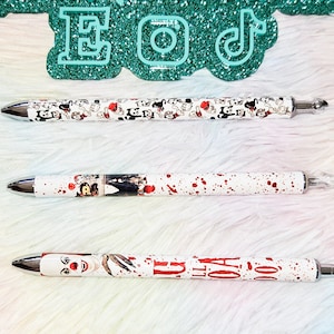 May include: Three pens with silver tips and white barrels. One pen features a pattern of cartoon characters, another has a horror movie theme with red splatters, and the third pen has a horror movie theme with the text "IT". The pens are displayed on a fluffy white surface.