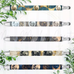 May include: Five stylus pens with moon phase designs. The pens have silver tips and black touch-screen ends. The pen barrels feature various moon phase patterns in shades of blue, gold, and black. Green foliage frames the pens.