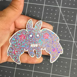 May include: A glittery moth sticker with the text "HUG?" in white. The moth has large, detailed wings with a purple and blue iridescent effect. The sticker is held against a gray cutting mat.