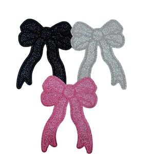 May include: Three glittery bow appliques in black, silver, and pink. Each bow has a detailed design with long, flowing ribbons. The bows are likely for crafts, decorations, or embellishments, adding a touch of sparkle to various projects.