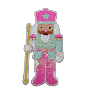 May include: A sequined Christmas nutcracker figure in pink, teal, and gold. The figure has a pink hat with a star, a teal jacket, pink boots, and a gold staff. The nutcracker has a white beard and a brown mustache.