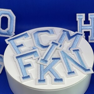 May include: A set of light blue felt letters with white outlines. The letters are F, C, M, K, and N. The letters are arranged in a pile on a white surface.