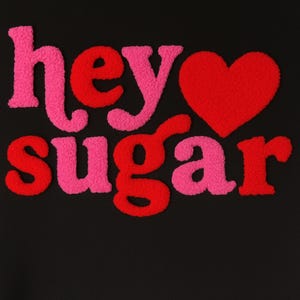 May include: Black background with the words "hey sugar" in pink and red, with a red heart. The letters have a fuzzy, textured appearance, creating a playful and eye-catching design. The overall aesthetic is bold and romantic.