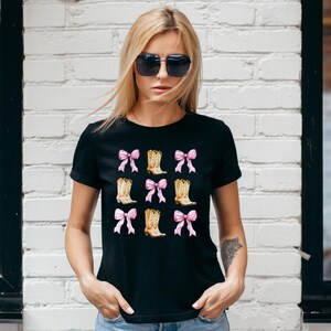 May include: Black t-shirt featuring a repeating pattern of pink bows and cowboy boots. The boots are tan with brown accents. The bows are a soft pink color. The shirt is a classic crew neck style.