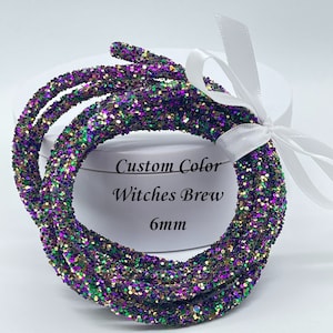 May include: A close-up of a spool of 6mm glitter cord in a custom color called "Witches Brew". The cord is a mix of green, purple, gold, and black glitter.