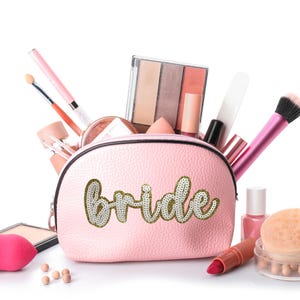 May include: Pink makeup bag with the word "bride" in gold glitter letters. The bag is open and filled with various makeup items, including brushes, eyeshadow palettes, and lipstick.