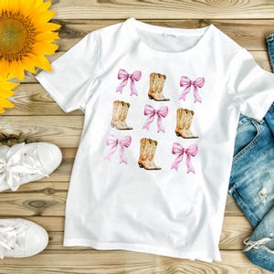 May include: White t-shirt featuring a repeating pattern of pink bows and cowboy boots. The boots are tan with brown accents. The bows are a soft pink color. The shirt is made of a soft, comfortable material.