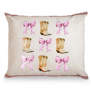 May include: A decorative pillow with a cream-colored background featuring a pattern of pink bows and cowboy boots. The boots are tan with intricate detailing. The pillow has a red striped border.
