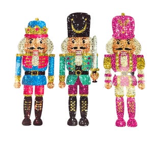 May include: Three decorative nutcrackers in a row, each with a unique color scheme. The nutcrackers have sparkling details, with blue, green, and pink outfits. Each nutcracker has a different hat and holds a sword or scepter.