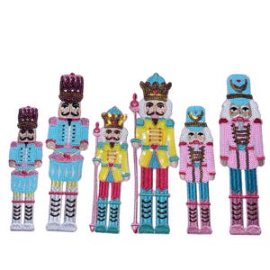 May include: A collection of six embroidered nutcracker figures in various sizes. Each figure features a detailed design with vibrant colors, including blue, pink, yellow, and red. They are adorned with sequins and beads, and have a regal appearance.