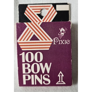 May include: A rectangular purple box containing 100 bow pins. The packaging features the text Pixie and 100 BOW PINS in white lettering. Above the text is a graphic of a stylized orange and white striped triangle with a circular hole for hanging.