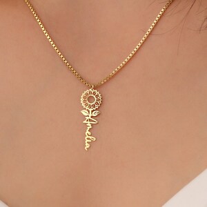 May include: A gold necklace with a sunflower pendant and the name "Amelia" written on the stem.